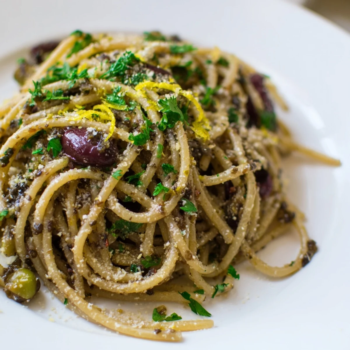 Garlicky olive tapenade pasta plated with lemon wedges, ready for a sprinkle of vegan Parmesan alternative.