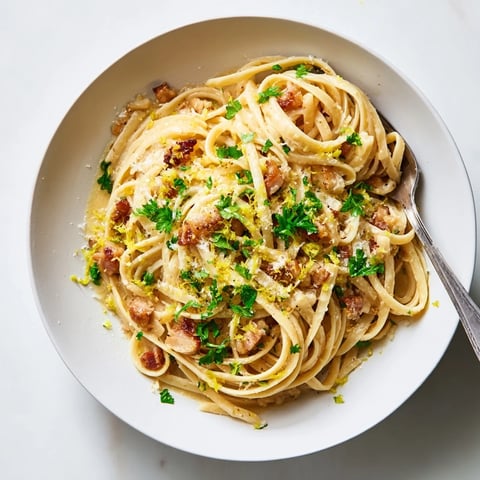 Golden-brown crispy bacon bits glisten atop freshly cooked linguine in a creamy, garlic-infused sauce, garnished with vibrant green parsley and lemon zest for a zesty finish.  