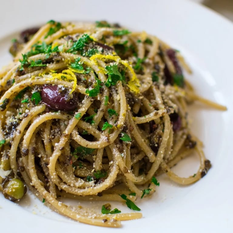 Garlicky olive tapenade pasta plated with lemon wedges, ready for a sprinkle of vegan Parmesan alternative.